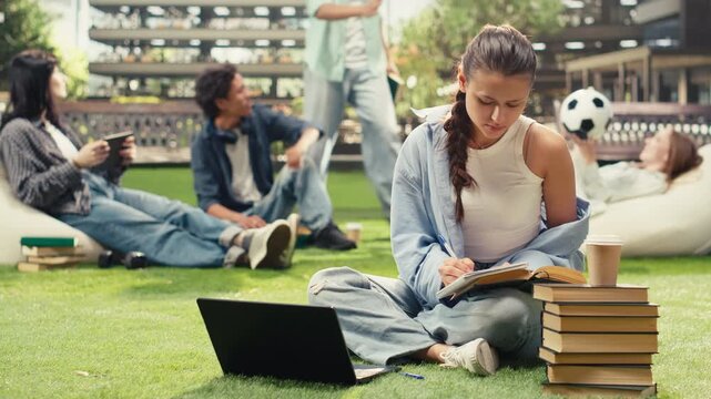 Focused student writes in a notebook beside an open book on a university campus lawn, taking coursework notes with stacked books and a backpack during an independent outdoor study session.