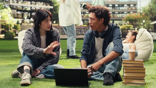 University students sit face to face on the grass with open textbook and stacked books, discussing study ideas during focused outdoor campus collaboration, supporting relaxed peer tutoring.