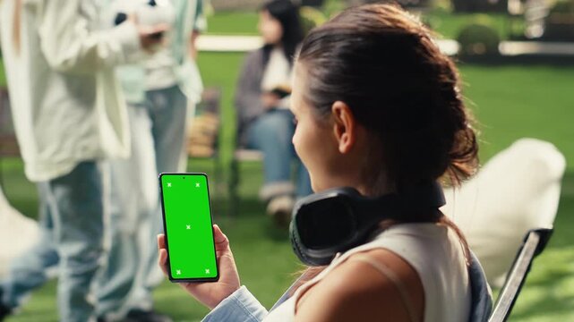 Student seated on a campus bench holds a smartphone with a vertical green screen, wearing headphones around her neck and presenting the editable mockup device in an outdoor academic environment.