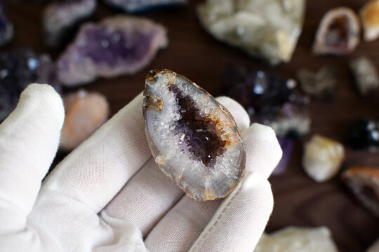 A hand in a white glove holds an agate geode with amethyst crystals against a background of gemstones