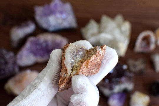 A white-gloved hand holds a stone with amethyst crystals on a rock against a background of precious stones.