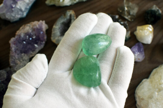 A hand in a white glove holds two stones of processed green fluorite against a background of gemstones