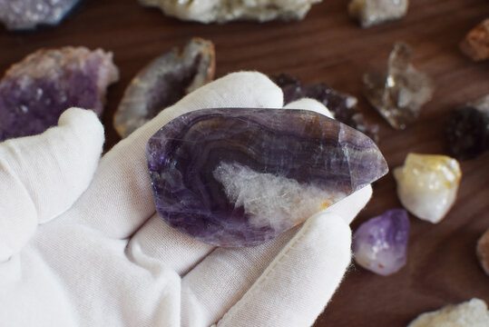 A white-gloved hand holds a polished striped purple fluorite against a background of gemstones