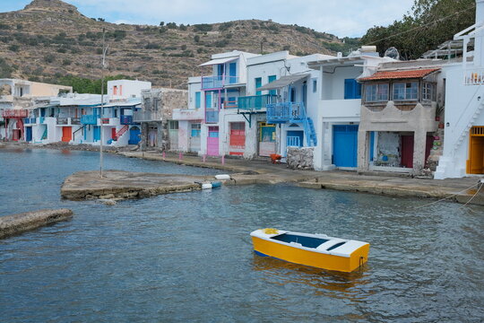 Colorful whitewashed fishing houses with blue, pink and yellow doors line the waterfront, with a bright yellow boat floating in the calm sea in Klima, Milos.
