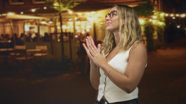 Woman in glasses clapping hands outside restaurant on street at night; appreciation evening moment.