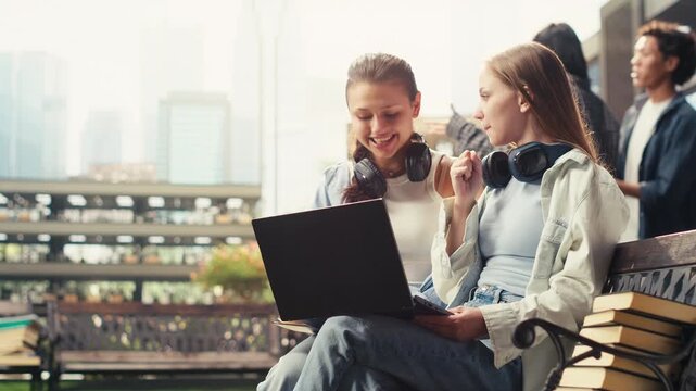 Women classmates collaborate on a laptop while seated on a bench at a university campus, discussing coursework and taking notes for exam preparation, supporting peer tutoring and group study.