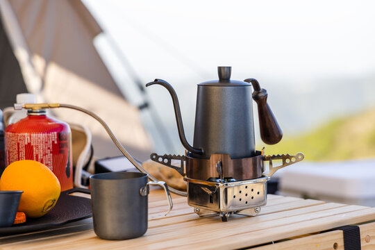 Portable camping coffee gear including a stove, kettle, cup and gas canister set up on a wooden table outdoors.