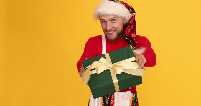 Happy man dressed as elf looking for somebody and giving Christmas gift on yellow background
