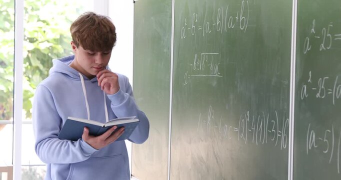 Teenage boy with book solving equation at chalkboard in classroom
