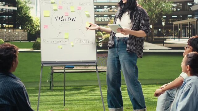 College student presents a strategy whiteboard with sticky notes as classmates listen at an outdoor campus tutoring session focused on project preparation, group study and academic collaboration.