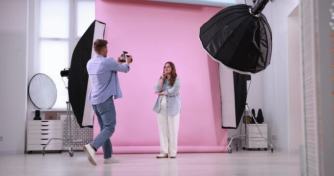 Plus size model posing for photographer with camera against pink background in studio. Beautiful young overweight woman