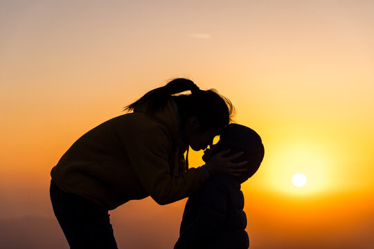 A silhouetted mother and child share an intimate, tender moment against a vibrant sunset sky, highlighting their close bond.