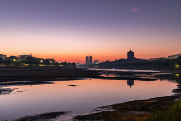 Fototapeta premium A serene riverside landscape at twilight, featuring a traditional tower, city buildings, and clear sky reflecting on calm water.