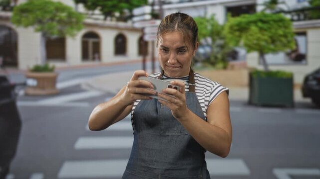 Woman holding smartphone tapping and swiping screen on street crosswalk with apron, focused expression; frustration concentration.