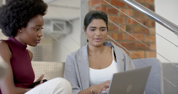 Diverse female coworkers sitting at office lounge, viewing silver laptop and phone, typing for work