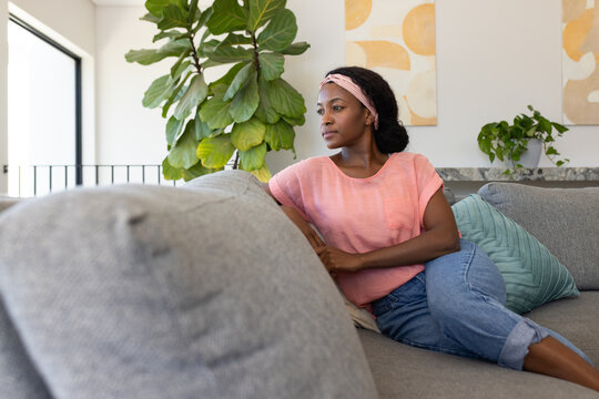 African woman reclining on grey sofa in living room, gazing toward balcony railing, copy space