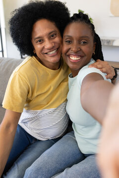 African American couple pressing cheeks and taking selfie on gray couch in living room with phone
