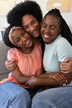 African American women sitting closely, hugging and smiling on grey sofa at home with wall art