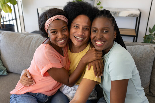 Diverse friends sitting close on grey couch in living room hugging and smiling at camera
