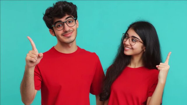 Two young people in red shirts point upward together. They wear glasses and smile at the camera. They stand against a solid teal background. Their gestures suggest excitement or discovery