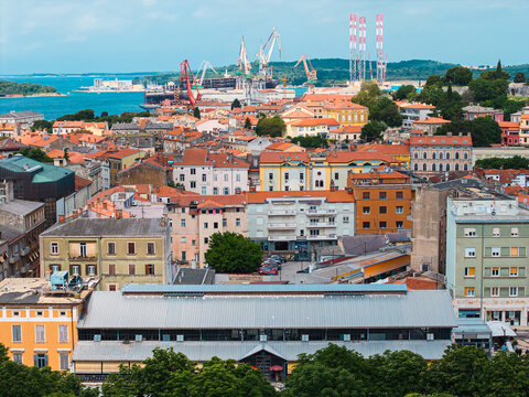 Aerial view of the historic city of Pula with its red-tiled rooftops, bustling harbor with industrial cranes, and the blue Adriatic Sea in Pula, Istria County, Croatia.