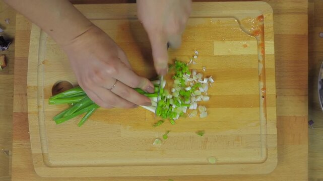 Woman cuts spring onion on wooden cutting board in kitchen during food preparation.