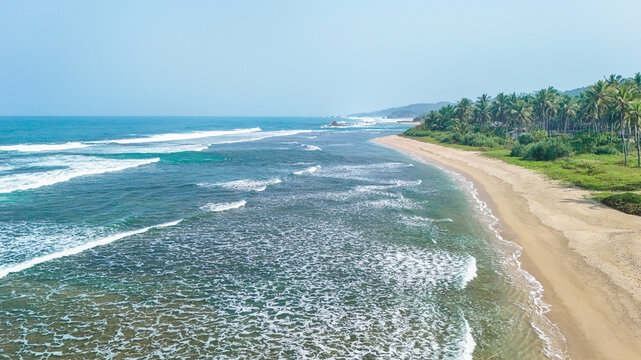 Aerial view of a tropical coastline with turquoise waves crashing onto a sandy beach, bordered by lush palm trees under a clear sky in Indonesia.