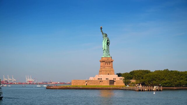 Statue of Liberty on Liberty Island in New York City. The copper monument rises above trees and harbor water with distant cranes on the horizon.