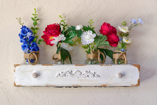 Silk flowers of various colors are displayed on the wall on a shelf made from an antique drawer
