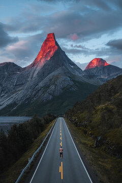 Aerial view of a lone person standing on a road leading toward the illuminated peak of Stetind mountain surrounded by dramatic mountains and water in Narvik Municipality, Nordland, Norway.