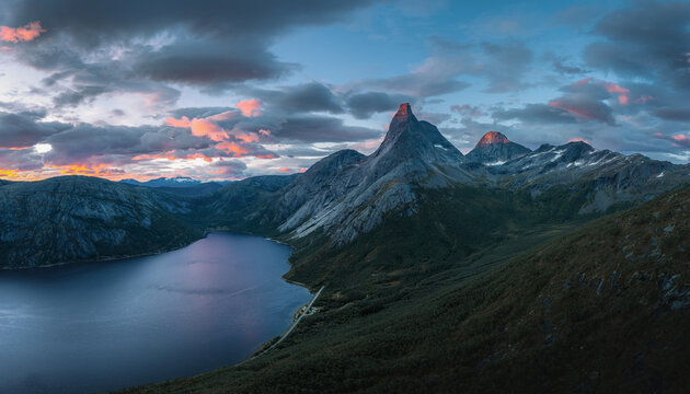 Aerial view of the iconic Stetind mountain rising above a deep blue lake with dramatic sunset clouds illuminating the sky in Narvik Municipality, Nordland, Norway.