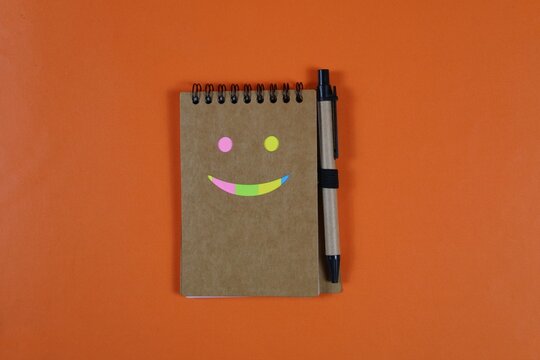Colorful smiley face made with sticky notes on a spiral notebook and pen, arranged on an orange background with ample copy space.