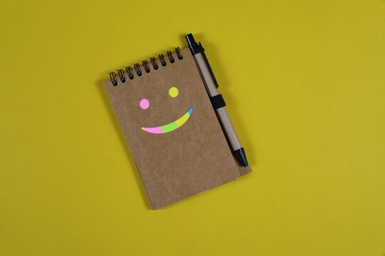 Colorful smiley face made with sticky notes on a spiral notebook and pen, arranged on an yellow background with ample copy space.