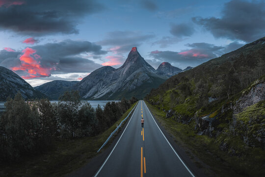Aerial view of Stetind mountain towering over a scenic road with a person walking in the distance under a dramatic sunset sky in Narvik Municipality, Nordland, Norway.