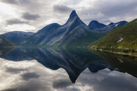 Aerial view of Stetind mountain reflected in the calm waters of the fjord under a cloudy sky in Narvik Municipality, Nordland, Norway.