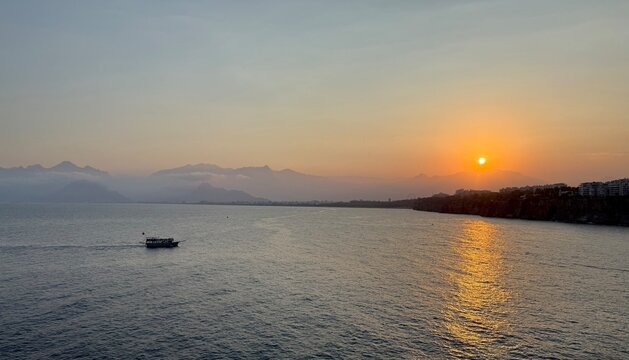 Excursion Boat On Mediterranean Sea At Sunset In Antalya. Wide shot of a small tourist ship sailing on calm sea water during golden hour sunset with the Taurus Mountains and rocky coastline