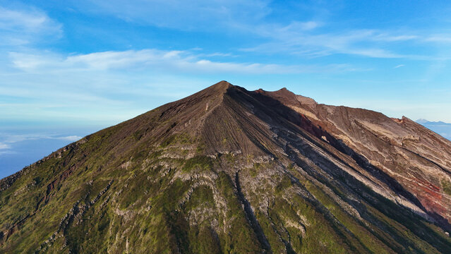 Aerial view of Mount Agung with its rugged volcanic slopes, rocky terrain, and patches of green vegetation under a clear blue sky in Rendang, Bali, Indonesia.