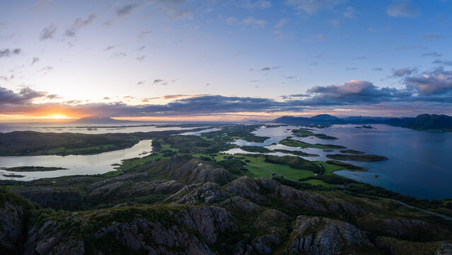 Aerial view of Torghatten mountain and surrounding coastal archipelago with rocky terrain, green fields, and calm waters under a vibrant sunset sky in Bronnoy Municipality, Nordland, Norway.