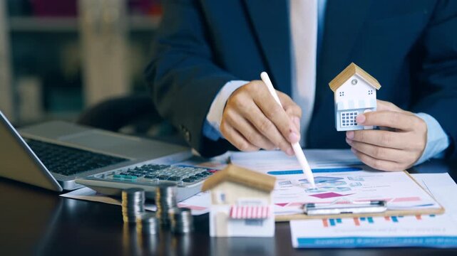 Real estate agent holding house model and using pen to evaluate property price and offer details for clients.