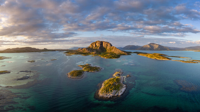 Aerial view of Torghatten mountain with its iconic hole, surrounded by the clear blue waters of the Norwegian Sea, rocky islands, and a soft sunset sky in Bronnoy, Nordland, Norway.