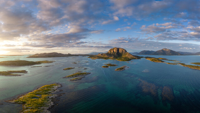 Aerial view of Torghatten mountain with its iconic natural tunnel, surrounded by coastal islands and clear blue waters under a dramatic cloudy sky in Bronnoy Municipality, Nordland, Norway.