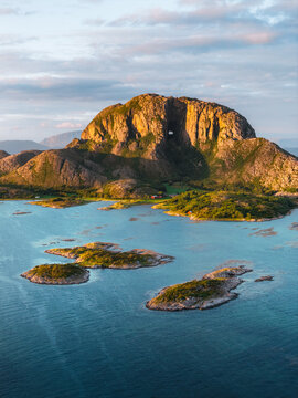 Aerial view of Torghatten mountain with its iconic natural tunnel, surrounded by small coastal islands and blue water under golden hour light in Bronnoy Municipality, Nordland, Norway.