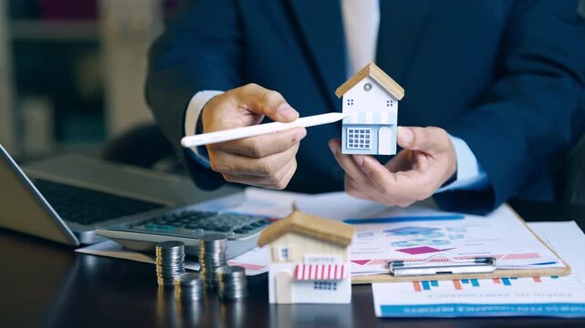 Real estate agent holding house model and using pen to evaluate property price and offer details for clients.