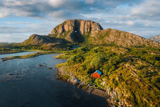 Aerial view of Torghatten mountain with its iconic hole, surrounded by lush greenery, rocky terrain, and a red wooden cabin near the coastline in Bronnoy Municipality, Nordland, Norway.