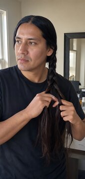 Handsome indigenous man braiding his long dark hair into a three strand braid standing in his bedroom