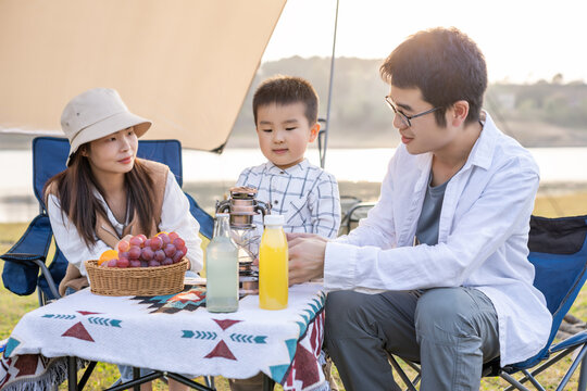 A family of three enjoys a picnic at an outdoor lakeside setup during daytime.
