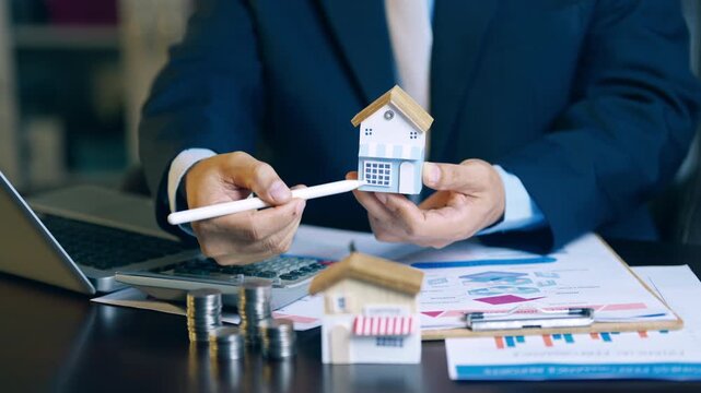 Real estate agent holding house model and using pen to evaluate property price and offer details for clients.