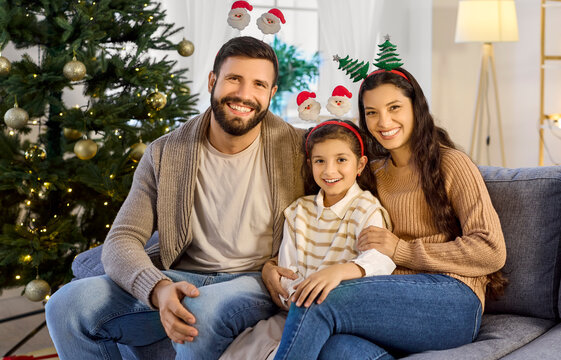 Christmas family portrait with smiling daughter at home. Parents and child girl sit on couch by festive tree, wearing cute headbands celebrating new year eve. Family love and winter holidays.