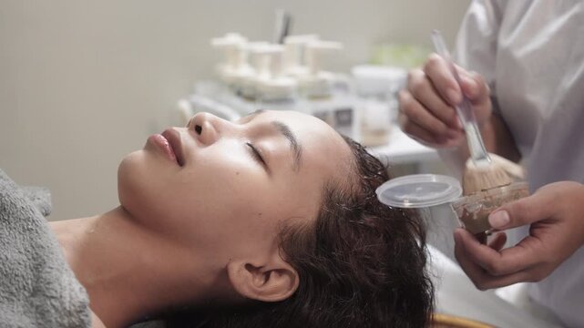 A southeast asian woman receiving a mineral mud clay facial mask application before a jade mask treatment at a Japanese head spa salon.