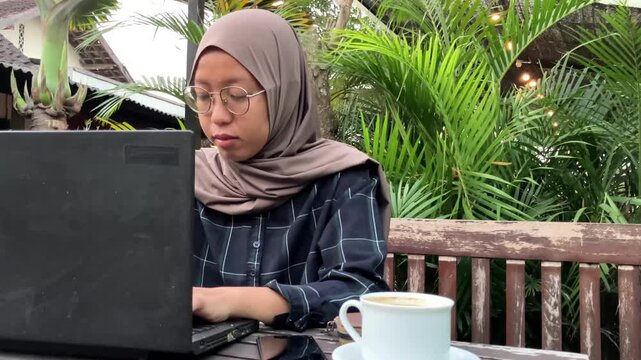 Slow motion video of a tired young Muslim woman wearing a hijab looking weary at her laptop screen. She sits at an outdoor cafe table with coffee. Remote freelance burnout and fatigue concept.
